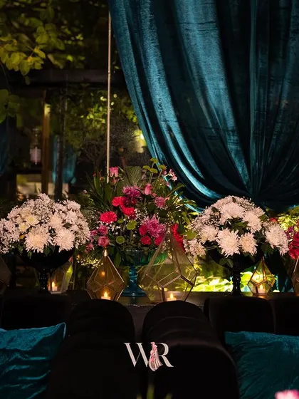 A close-up of the floral arrangements behind a plush black sofa with teal cushions. The mix of pink, white, and deep red flowers adds a pop of color to the moody decor.