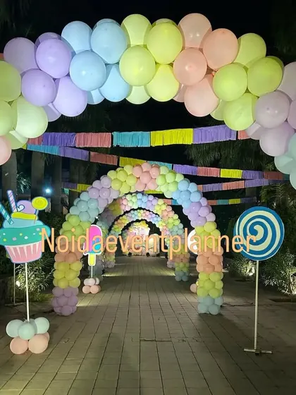 An immersive candyland party entrance featuring a long tunnel of pastel rainbow balloon arches and cute lollipop and cupcake props along the walkway.