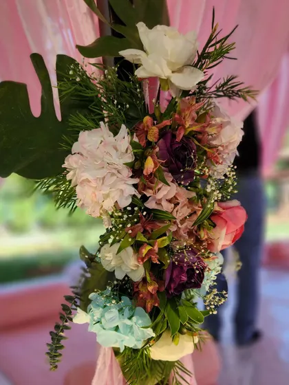 A close-up of the florals used to adorn a mandap post. This arrangement features a vibrant mix of flowers in shades of purple, pink, and light blue, accented with a large monstera leaf for a touch of tropical flair.