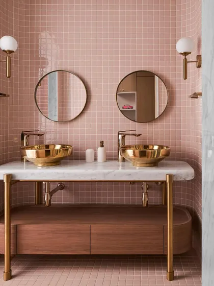The kid's bathroom features a playful yet chic design with three shades of pink tiles. A double vanity with bold brass sinks and fittings provides a functional and stylish space for two.