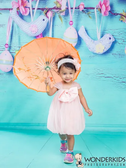 A sweet toddler in a pink dress holds a small umbrella under hanging bird and egg decorations. The bright turquoise background and spring-themed props create a cheerful and colorful photo.