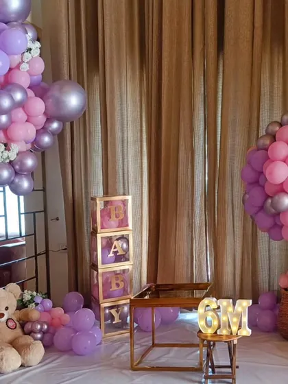 Celebrating a half-birthday is a growing trend. This indoor setup for a six-month milestone features purple and pink balloon arrangements, teddy bears, and a light-up '6M' sign.