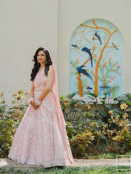 A full-length portrait of the bride in her elegant pink lehenga, standing in a beautiful garden setting.