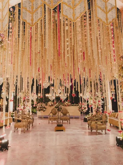 A wide shot of the magnificent floral mandap, showcasing the intricate ceiling work with hanging tassels and chandeliers, and the plush seating arrangement for guests.