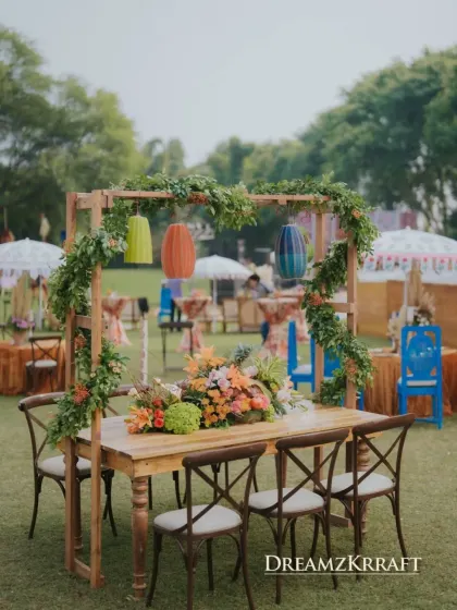 A beautiful dining setup at the Marrakesh Mehendi, with a wooden pergola draped in greenery and colorful lanterns. The cross-back chairs add a touch of rustic charm.