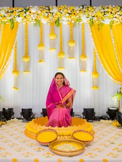 A beautiful portrait of the bride seated against the vibrant yellow and white decor of her haldi ceremony stage.