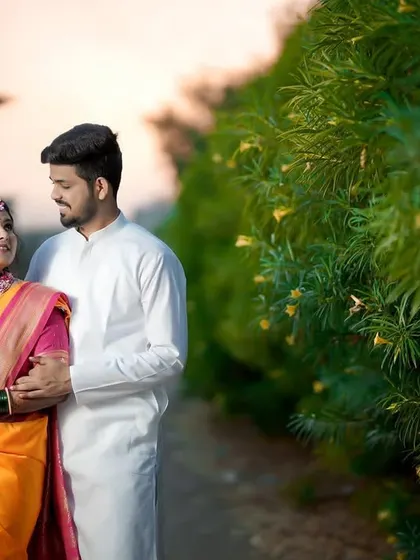 A wide shot of the couple standing together in a natural, outdoor setting. The soft evening light adds a romantic glow to this portrait from their Haldi function.