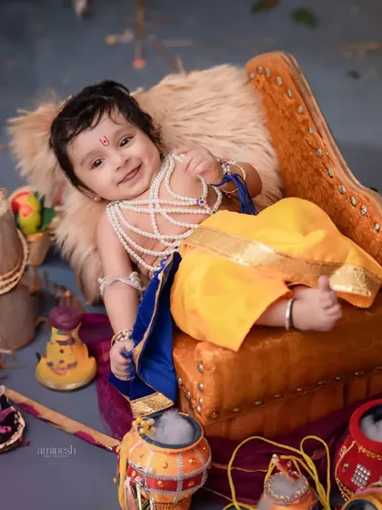 A happy baby dressed as Krishna, relaxing on a small couch surrounded by festive props. This is a fun, casual shot that captures the joy of the theme.