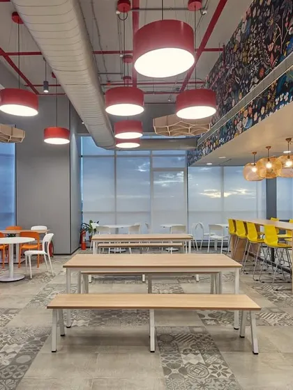 A large, bright cafeteria with a variety of seating options, including long communal tables, smaller round tables, and a high-top counter. The space is designed to accommodate a large number of people comfortably.