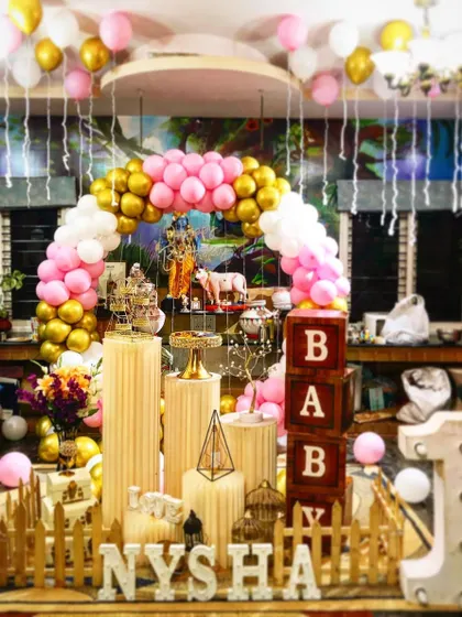 A first birthday and naming ceremony setup with pink, white, and gold balloons. The decoration includes 'BABY' blocks and marquee letters for the name.