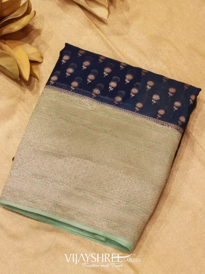 A dark blue Kora silk saree with a large silver zari border and delicate floral motifs. Kora silk is known for its texture and strength.