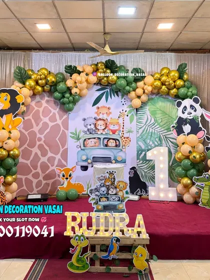 A full-stage jungle animal theme for Rudra's first birthday. The setup includes a large backdrop, multiple animal cutouts, a balloon arch, and a custom wooden name sign.