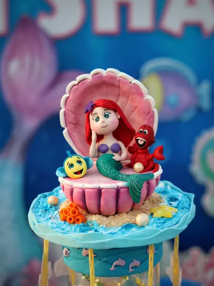 A stunning Little Mermaid cake topper, perfect for an under-the-sea party. This piece features Ariel, Flounder, and Sebastian nestled inside a large, detailed clamshell.