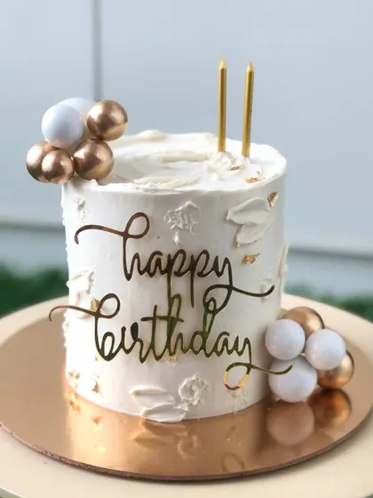 A simple and elegant white birthday cake with gold accents and a modern script topper.