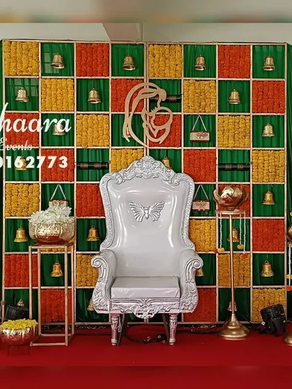 A traditional baby shower stage with a unique backdrop. The grid features squares of marigolds and hanging bells, with a mother-and-child motif, blending classic elements with a modern design.