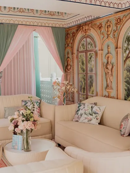 A comfortable lounge area with cream sofas and floral cushions. The walls are painted to resemble a romantic, sunlit conservatory, and the pastel pink and green drapery adds to the soft, dreamy atmosphere.