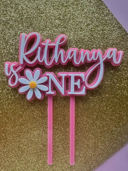 A sweet daisy-themed first birthday topper for "Rithanya." The layered pink and white acrylic design is fresh, modern, and perfect for a little one's celebration.
