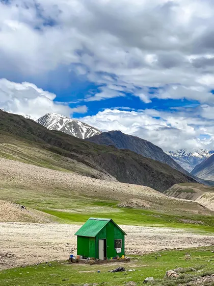 A small, green hut stands alone in a vast, barren landscape in Spiti Valley. This image highlights the solitude and immense scale of the Himalayan wilderness.