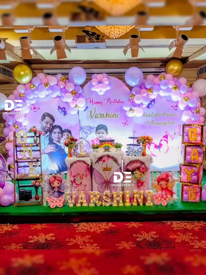 A personalized butterfly theme for Varshini's first birthday. The backdrop includes a lovely family photo, surrounded by purple balloons, butterfly cutouts, and light-up letters, making it extra special.