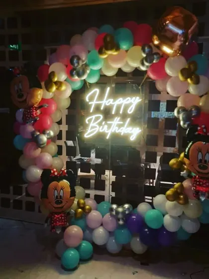 A classic Minnie Mouse themed birthday party. A circular balloon arch in pastel colors is decorated with Minnie Mouse foil balloons and a central 'Happy Birthday' neon sign.
