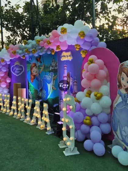 A dreamy Sofia the First princess party setup on a lush green lawn. The backdrop features the princess herself, a castle, and a beautiful balloon arch in shades of purple, pink, and gold.