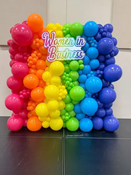 A vibrant rainbow balloon wall with a 'Women in Business' neon sign, created for a corporate event or conference.