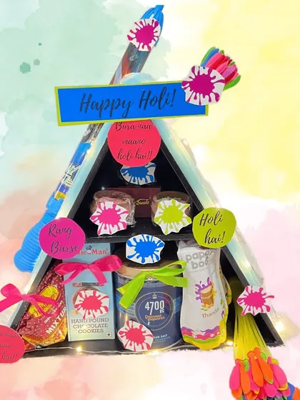 A triangular 'Happy Holi' hamper filled with treats and festive items like pichkaris and water balloons.