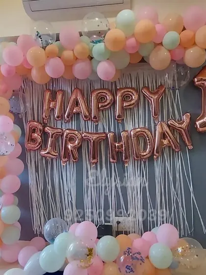 A lovely pastel balloon wall decoration for a first birthday at home. The setup includes a white fringe curtain, rose gold foil letters, and a frame of pastel balloons with confetti accents.