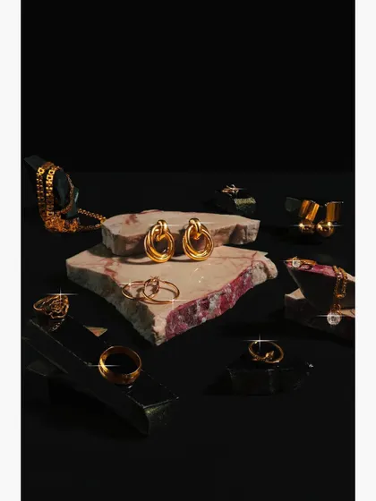 A collection of gold jewelry, including rings and earrings, artfully arranged on pieces of raw marble. The dark, moody lighting and contrasting textures create a luxurious and high-end feel for a brand catalog.