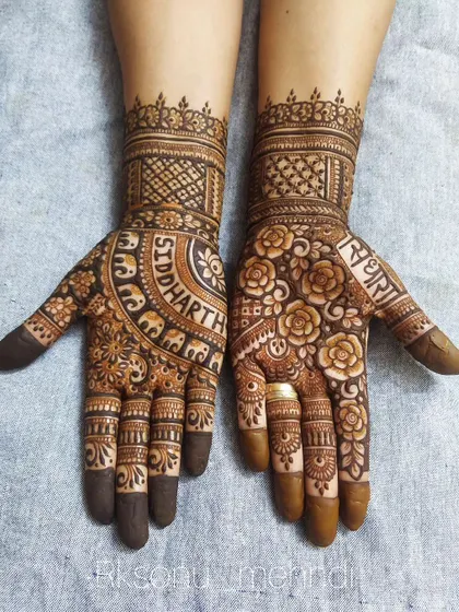 A unique design where the groom's name, "Siddharth," is written in a bold arc on one hand, surrounded by elephant motifs. The other hand features a dense floral pattern with the bride's name, "Radha."