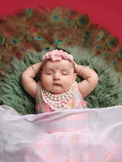 A beautiful close-up on the face of the sleeping 'Little Radha', highlighting her delicate features and the pearl necklace against the backdrop of peacock feathers.