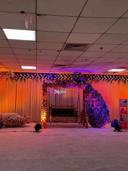 This wide-angle shot shows a complete baby shower stage design in a large hall. The setup includes a floral moon cradle, a decorated jhula swing, and a large balloon backdrop, all arranged on a white carpeted stage with spotlights.