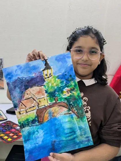 This student proudly displays her vibrant watercolor landscape of a European town, showcasing her use of bold colors and expressive brushstrokes.