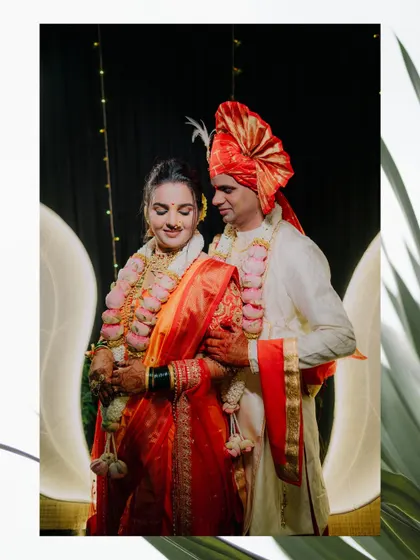 Purva and Dharma framed by elegant decor. The bride's red and orange saree contrasts beautifully with the groom's white sherwani and red pheta.