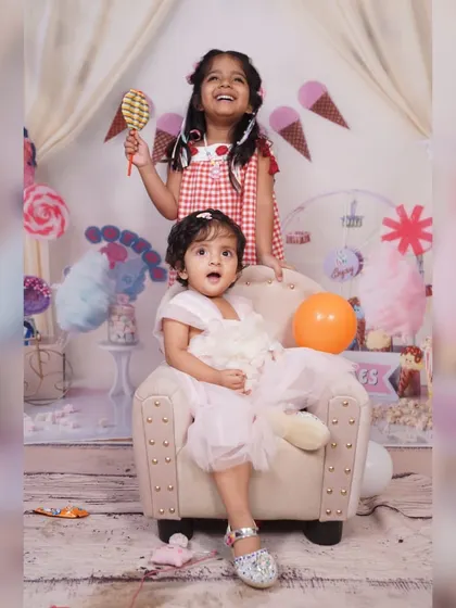 Sibling love and laughter. The older sister stands behind her baby sister, who is seated in the mini armchair, creating a fun and interactive portrait during the photoshoot.