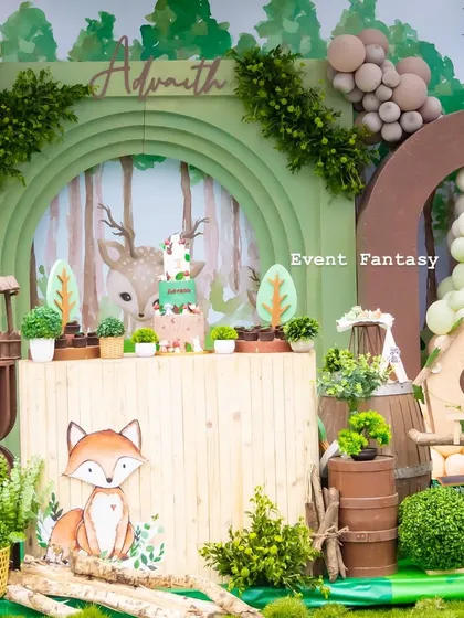 A beautiful woodland cake table for Advaith. The setup features a green arched backdrop with a forest illustration, a cute fox cutout, and various rustic elements like a wishing well and birdhouse.