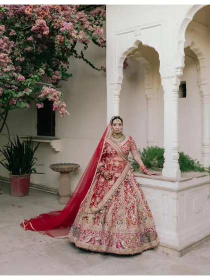 A classic bridal portrait set against the iconic arches and bougainvillea of a Jaipur palace. The vibrant red lehenga contrasts beautifully with the timeless architecture.