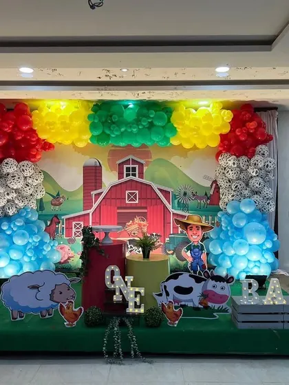 A charming Farm Animal theme for a first birthday. The backdrop features a big red barn, with cutouts of a farmer and friendly farm animals, surrounded by colorful balloon clusters.