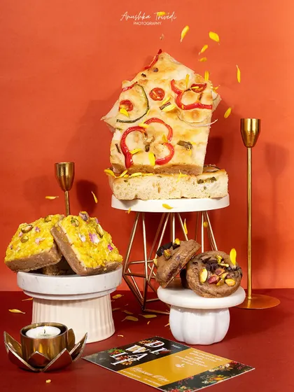 An artistic, elevated shot of focaccia bread and other Diwali treats, styled with falling petals for a dynamic effect.
