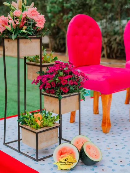 A vibrant pink chair stands out against the fresh green grass, with tiered planters holding colourful flowers and fresh watermelons adding a playful touch.
