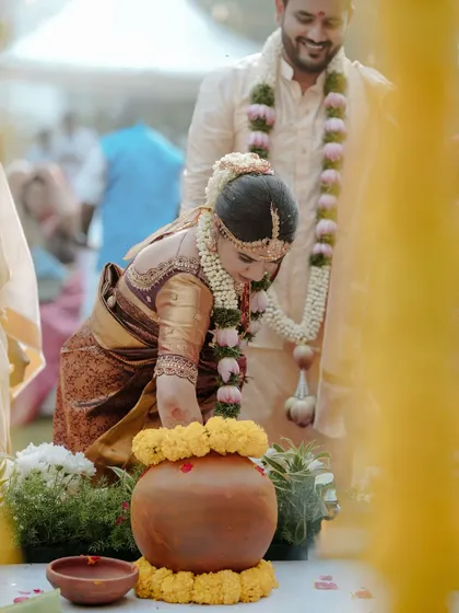 A candid shot of a bride performing a ritual during her South Indian wedding ceremony, with the groom smiling in the background.