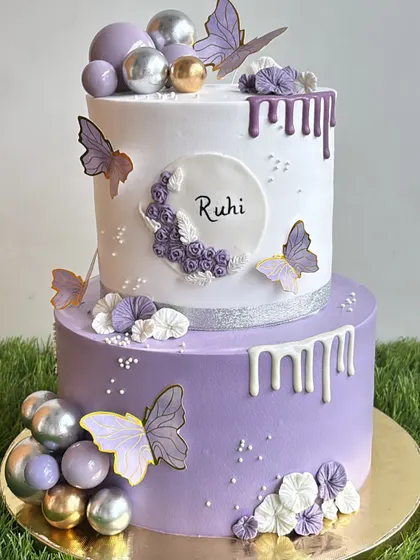 This elegant two-tiered birthday cake combines soft violet and white whipped cream frosting. We decorated it with delicate butterflies, chocolate drips, and shimmering spheres for a sophisticated and beautiful celebration.