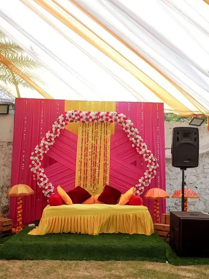 A bright and cheerful Haldi setup for a destination wedding. The decor features a pink and yellow backdrop with a hexagonal floral frame and personalized photos of the couple.