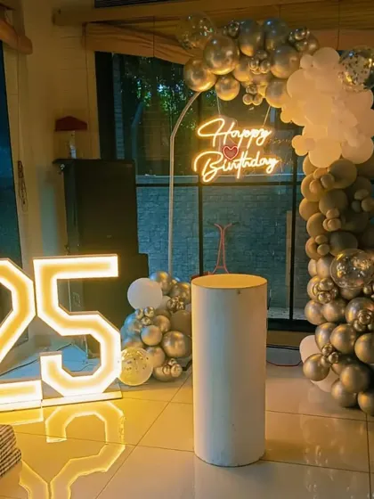 A 25th birthday party with a silver and white balloon arch. This setup includes a "Happy Birthday" neon sign and large marquee numbers.