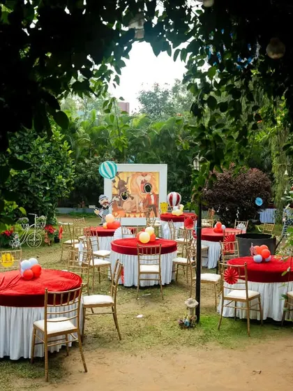 An outdoor carnival setup in a beautiful garden. The round tables are covered with red tablecloths, and the area is decorated with balloons and themed props.