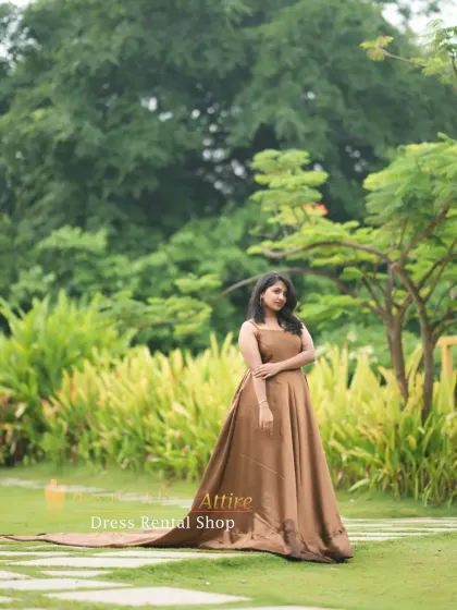 A full-length portrait featuring the gold satin trail gown in a garden. The simple, classic cut of the dress is elevated by the dramatic, long tail.