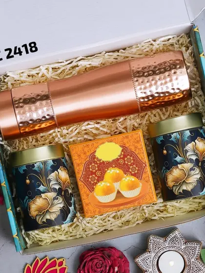 A luxurious Diwali gift box featuring a hammered copper bottle, two decorative tins for sweets or dry fruits, and a box of motichoor ladoo. The set is completed with festive diyas for the occasion.
