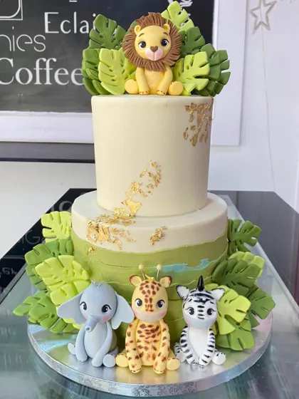 A sophisticated two-tiered safari cake with gold leaf accents. This design features cute, stylized fondant animals including a lion, elephant, giraffe, and zebra.