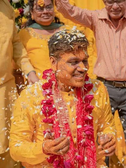 A portrait of the groom smiling, covered in turmeric and flower petals, with a garland of red flowers.