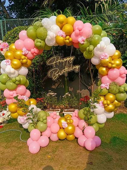 An outdoor balloon ring decoration in a garden. The pink, green, and gold balloons are mixed with small floral accents for a fresh, natural look.